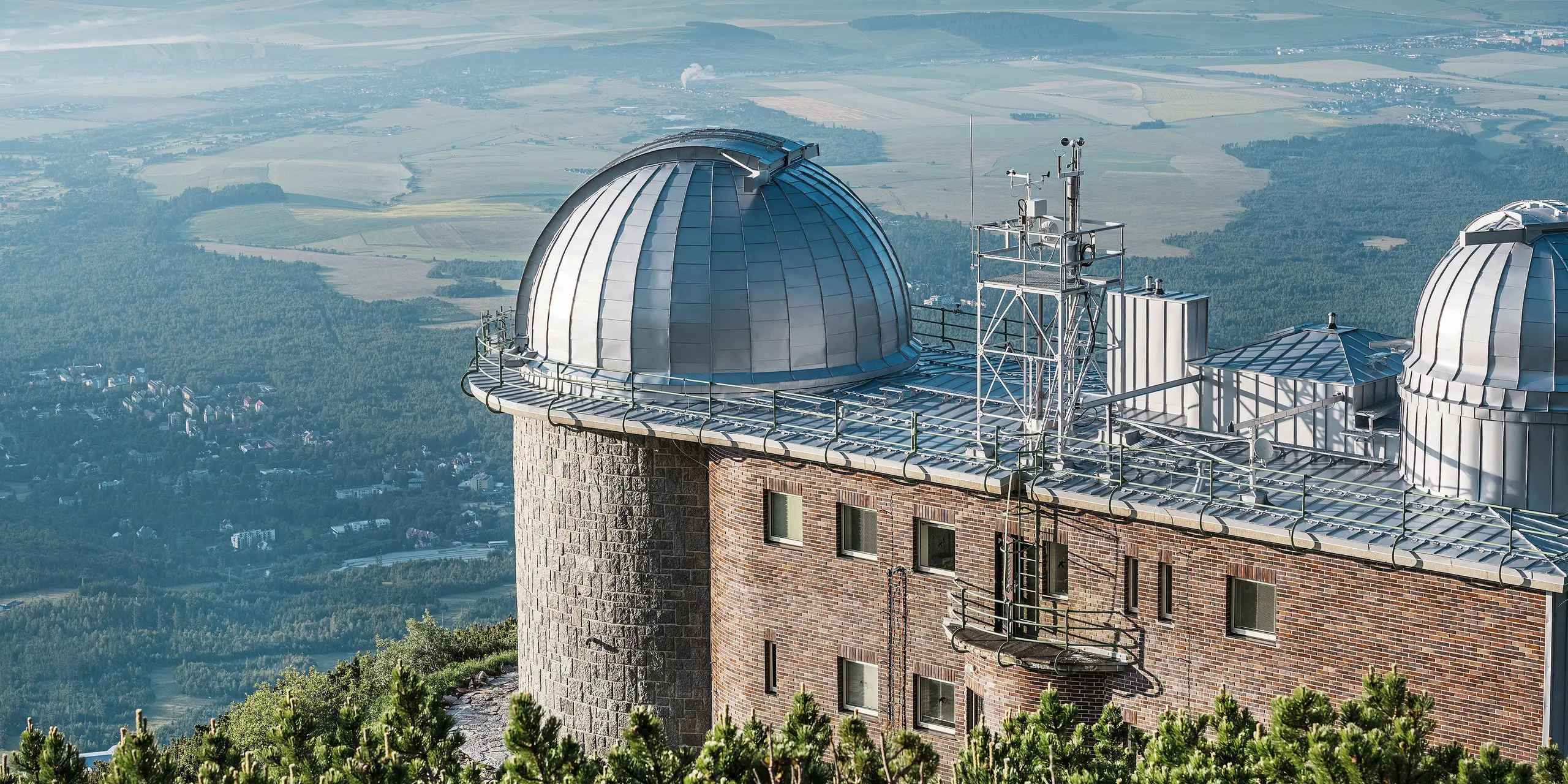 Observatorium Skalnaté Pleso, über der idyllischen slowakischen Landschaft gelegen, mit glänzenden PREFALZ Kuppeln in Silbermetallic. Das imposante Gebäude fügt sich nahtlos in seine natürliche Umgebung ein und thront majestätisch auf einem Bergkamm. Dank der langlebigen PREFA Aluminiumverkleidung, die auch extremen Witterungsbedingungen standhält, bleibt die historische Sternwarte Tag und Nacht ein Hüter der Sterne. Das robuste und langlebige Material PREFA unterstreicht die Verpflichtung zu Qualität und Nachhaltigkeit, während die Architektur eine Brücke zwischen Himmel und Erde schlägt.