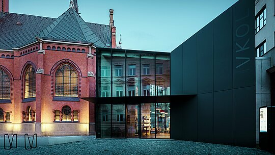 Slika prikazuje impresivnu fuziju povijesne i moderne arhitekture u Olomoucu. Istaknuta Crvena crkva sa svojim gotičkim stilom, uključujući umjetnički oblikovane prozore i visoki toranj, nadopunjena je modernim novim objektom s minimalističkom, crnom fasadom od PREFABOND aluminijsko-kompozitnih ploča. Prozračne staklene površine novog objekta reflektiraju povijesno okruženje i stvaraju fascinantan kontrast s čvrstom strukturom crkve. Precizna obrada modernih materijala, u kombinaciji sa skladnim povezivanjem starog i novog, daje zgradi suvremenu eleganciju, istovremeno poštujući povijest okoline. Ova kombinacija inovativnih i dugovječnih materijala poput PREFABOND ploča osigurava i estetsku ljepotu i funkcionalnost.