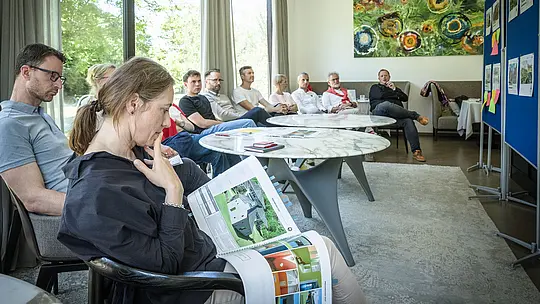 PREFARENZEN 2027 – stručna rasprava u svijetloj prostoriji za seminare: Paulina Frey (voditeljica marketinga PREFA CH) drži veliku arhitektonsku knjigu s projektima izrađenima od PREFA aluminija, dok ostali članovi žirija pažljivo slušaju. Veliki prozori propuštaju dnevno svjetlo i pružaju pogled na zeleno vanjsko okruženje. Scena prenosi dojam usredotočene analize, kreativne rasprave te vrednovanja dizajna i funkcionalnosti. PREFA aluminijski proizvodi pritom simboliziraju dugotrajne, estetski oblikovane krovove i fasade koje u arhitektonskim projektima potiču inovaciju, kvalitetu i inspiraciju.