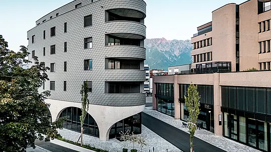 Pogled na Bernerhaus u Buchsu s upečatljivim trokutastim oblikom i nježno zaobljenim balkonima na pozadini alpskog krajolika. Kontinuirana fasada od PREFA zidnih šindra u P.10 svijetlosivoj boji obavija zgradu poput ljuskaste aluminijske kože i stvara živopisnu igru svjetla i sjene. Iznad masivnog betonskog postolja s elegantnim lukovima, lagana, otporna na vremenske uvjete PREFA aluminijska fasada djeluje posebno precizno, dugotrajno i gotovo bezdržavno – arhitektonski privlačan element u urbanom okruženju.