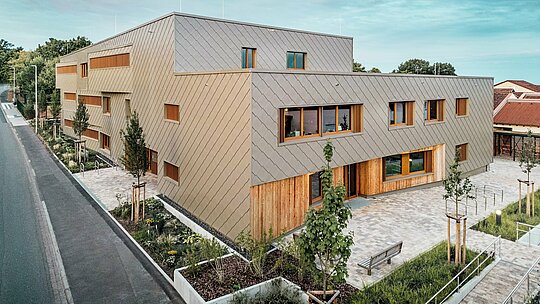 Večernja atmosfera obasjava dječji vrtić u Alzenauu nježnim svjetlom i ističe visokokvalitetnu fasadu od brončano obojenih PREFA zidnih rombova 44 × 44. Kombinacija strukturirane aluminijske fasade i vertikalnih drvenih površina stvara živahnu igru materijala, boja i tekstura. Prostrani prozori osiguravaju svijetle unutarnje prostore, dok okolne zelene površine i popločani predprostor pozivaju na boravak. Slika dojmljivo pokazuje kako se suvremena arhitektura, funkcionalnost i održivost mogu skladno povezati uz PREFA proizvode – usred gradskog konteksta Alzenaua.