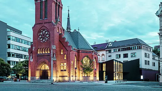 Slika prikazuje skladnu fuziju povijesne i moderne arhitekture u Olomoucu. Impresivna Crvena crkva, s upečatljivim šiljastim tornjem i gotičkim prozorima, čini povijesno središte ansambla. Odmah do nje uzdiže se moderni novi objekt čija fasada od matiranih PREFABOND aluminijsko-kompozitnih ploča i stakla stvara jasan, elegantan kontrast tradicionalnoj arhitekturi. Velike staklene površine omogućuju pogled u unutrašnjost novog objekta, koji je inovativno povezan s crkvom. Moderna zgrada, sa svojom preciznom formom i čistim linijama, doprinosi suvremenoj atmosferi, a da pritom ne narušava povijesni karakter crkve. Ova uspješna simbioza pokazuje kako visokokvalitetno obrađeni materijali poput PREFABOND ploča pružaju savršenu osnovu za spajanje tradicije i moderne arhitekture.