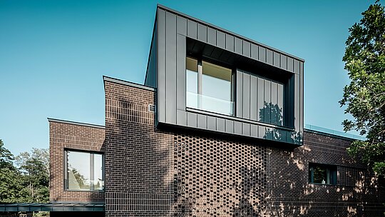 Snimka prikazuje upečatljiv fasadni dizajn moderne višegeneracijske vile u Budimpešti, pri čemu PREFALZ aluminij u boji P.10 antracit postavlja jasan arhitektonski naglasak. Istaknuti kubični volumen s preciznim stojećim falcevima stvara snažan kontrast strukturiranoj fasadi od opeke ispod njega. Bočno sunčevo svjetlo oblikuje dinamičnu igru sjena i linija koja naglašava visokokvalitetnu izradu i dugotrajnost PREFA aluminijskog rješenja. Kombinacija metala i klinkera dodatno ističe moderan, kontrastni karakter zgrade.