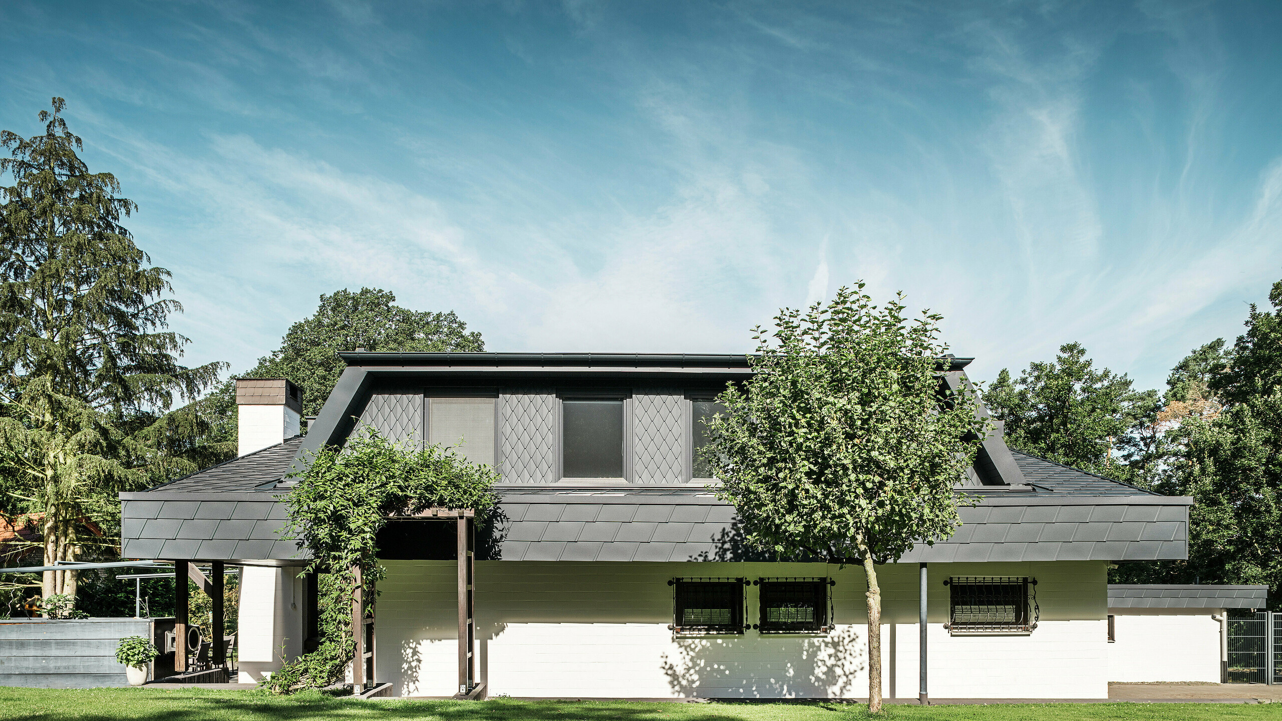 Pogled sprijeda na vrtnu stranu samostojeće kuće u Mechernichu, s velikim trapezastim krovnim prozorom obloženim malim PREFA P.10 krovnim crijepovima u antracitnoj boji. Krov je prekriven PREFA DS.19 krovnim šindrama u istoj boji. Fasada prizemlja obojena je u bijelo, a zelena pergola upotpunjuje cjelokupni izgled u vrtnom prostoru. Drveće i grmlje uokviruju pogled i osiguravaju skladnu integraciju u prirodno okruženje.