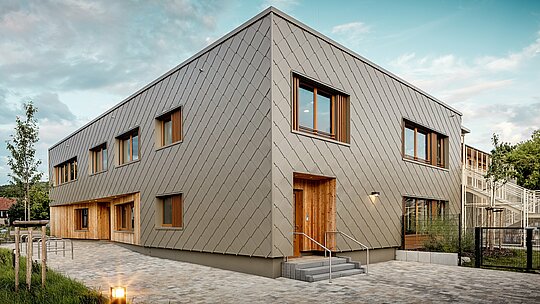 Moderni dječji vrtić u Alzenauu impresionira svojom jasnom arhitekturom i visokokvalitetnom PREFA aluminijskom fasadom u brončanoj boji. Fasada od zidnih rombova 44 × 44 daje zgradi elegantan, strukturiran izgled i vizualno povezuje različite funkcionalne prostore u skladnu cjelinu. Drvene obloge na ulazima stvaraju topao, prirodan kontrast dugotrajnom aluminiju. Kombinacija masivne konstrukcije, drvenih elemenata i ventilirane, prednje postavljene fasade primjer je održive gradnje. Održiva PREFA „odora“ zgrade impresionira ne samo reciklabilnošću, već i otpornošću na vremenske uvjete – idealno za budućnosno orijentirane obrazovne objekte s visokim arhitektonskim standardima.