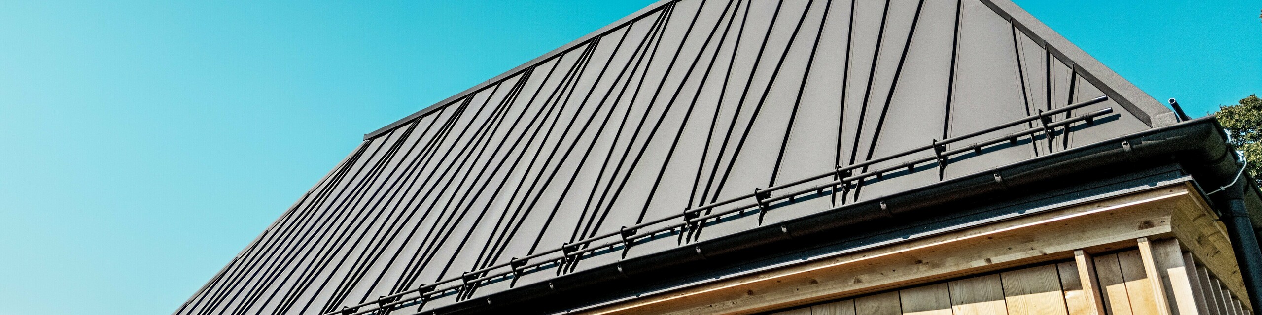 Krupni plan izvanrednog PREFA krova s falcom „Badehausa“ pri hotelu Hirschen u Schwarzenbergu. Krov u boji P.10 orah smeđa ističe se svojim upečatljivim, dijelom dijagonalno postavljenim falcima, koji zgradi daju dinamičan, moderan izgled. Jasna linija krova i nagib od 60 stupnjeva stvaraju snažan geometrijski dojam. Ispod krova vidi se fasada s vertikalno postavljenim drvenim lamelama, koje na nov način interpretiraju tradicionalnu gradnju. Bezoblačno nebo i direktno sunčevo svjetlo naglašavaju visokokvalitetnu površinu PREFALZ materijala i precizan majstorski rad. Fotografija dojmljivo prikazuje spoj inovacije, zanatstva i regionalnog stila.