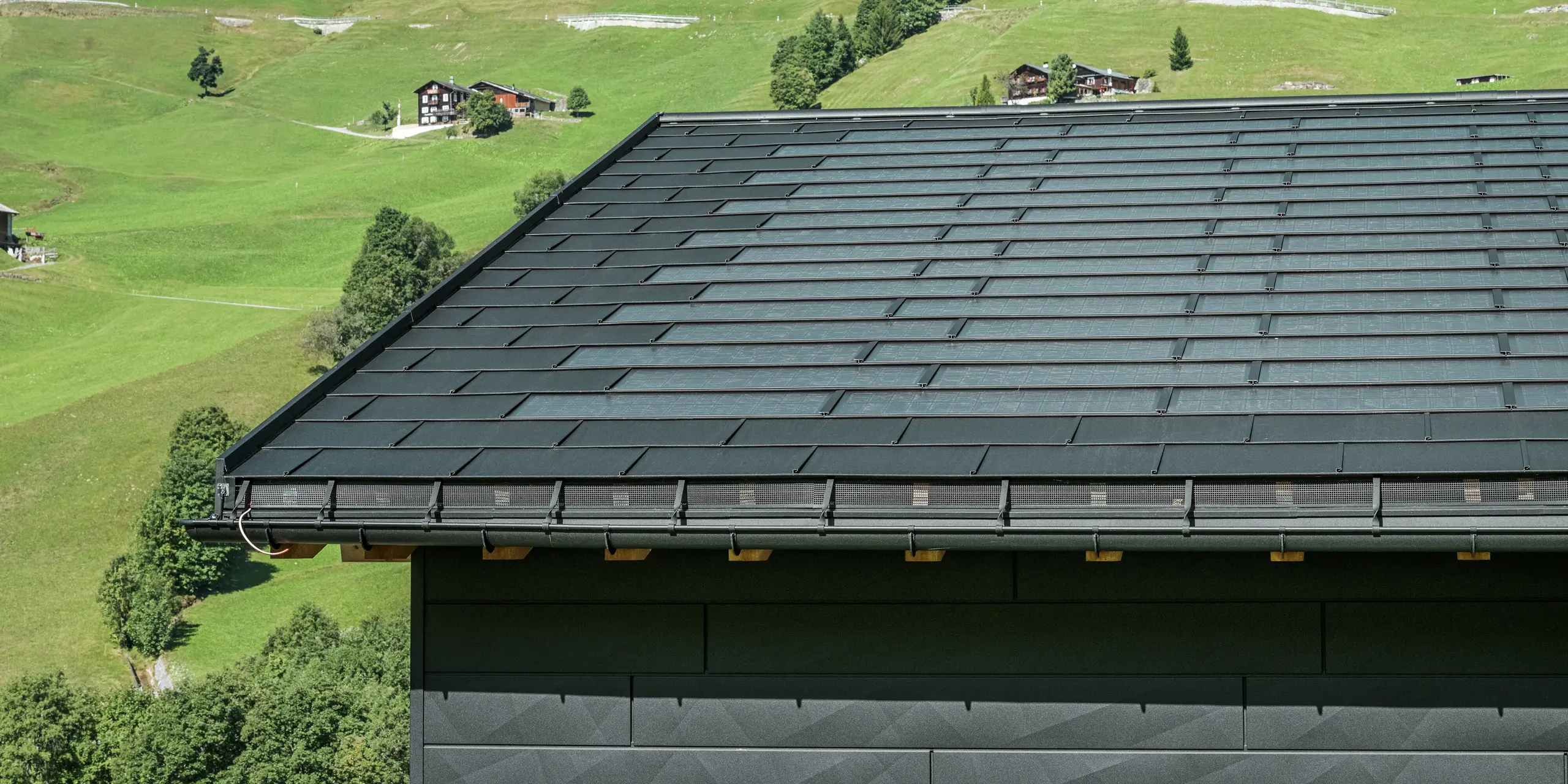 Krupni plan strehe moderne obiteljske kuće u alpskom okruženju Ascharine u Švicarskoj. Krov je pokriven inovativnim PREFA solarnim krovnim panelom i tradicionalnim krovnim panelom R.16, oba u boji P.10 Black. PREFA viseći oluk i PREFA odvodna cijev osiguravaju pouzdanu odvodnju krova. Prepoznatljive fasadne ploče PREFA Siding.X mogu se vidjeti na dnu slike. Čiste linije aluminijskih proizvoda naglašavaju moderan dizajn usred slikovitog planinskog krajolika.