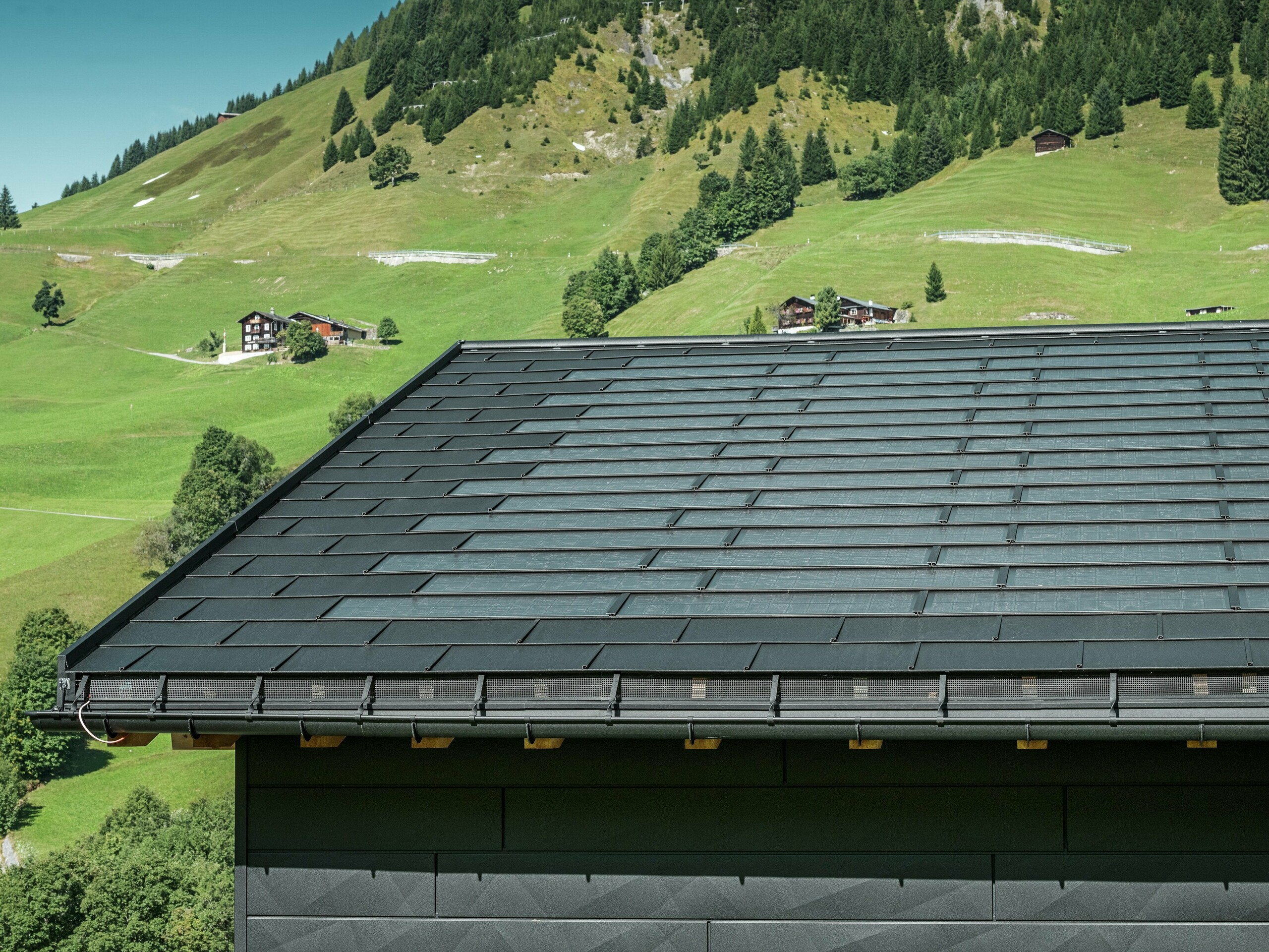 Krupni plan strehe moderne obiteljske kuće u alpskom okruženju Ascharine u Švicarskoj. Krov je pokriven inovativnim PREFA solarnim krovnim panelom i tradicionalnim krovnim panelom R.16, oba u boji P.10 Black. PREFA viseći oluk i PREFA odvodna cijev osiguravaju pouzdanu odvodnju krova. Prepoznatljive fasadne ploče PREFA Siding.X mogu se vidjeti na dnu slike. Čiste linije aluminijskih proizvoda naglašavaju moderan dizajn usred slikovitog planinskog krajolika.