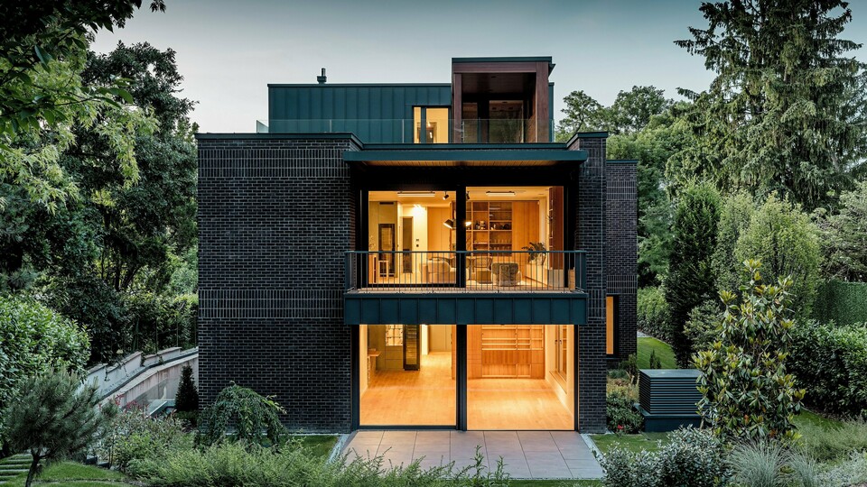 Das Bild zeigt die moderne Rückseite einer Mehrgenerationenvilla in Budapest in der Abenddämmerung. Die großen Glasflächen geben den Blick auf die warm beleuchteten Innenräume frei, während die Kombination aus dunkelroter Backsteinfassade und PREFALZ-Aluminium in der Farbe P.10 Anthrazit die klaren architektonischen Linien betont. Die präzise ausgeführten Stehfalze unterstreichen die hohe Verarbeitungsqualität und Langlebigkeit der PREFA-Aluminiumprodukte. Umgeben von üppigem Grün fügt sich das Gebäude harmonisch in die Gartenlandschaft ein und strahlt einen eleganten, modernen Charakter aus.