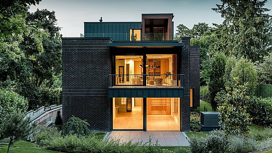 Das Bild zeigt die moderne Rückseite einer Mehrgenerationenvilla in Budapest in der Abenddämmerung. Die großen Glasflächen geben den Blick auf die warm beleuchteten Innenräume frei, während die Kombination aus dunkelroter Backsteinfassade und PREFALZ-Aluminium in der Farbe P.10 Anthrazit die klaren architektonischen Linien betont. Die präzise ausgeführten Stehfalze unterstreichen die hohe Verarbeitungsqualität und Langlebigkeit der PREFA-Aluminiumprodukte. Umgeben von üppigem Grün fügt sich das Gebäude harmonisch in die Gartenlandschaft ein und strahlt einen eleganten, modernen Charakter aus.