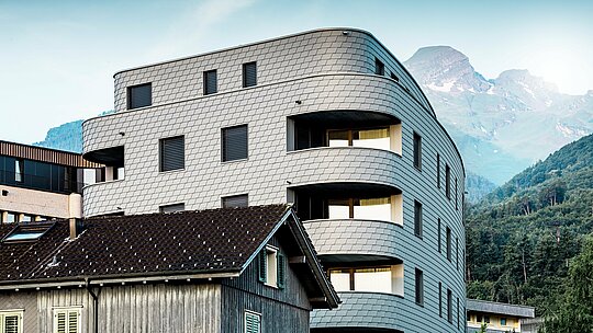 Moderna, trokutasta stambeno-poslovna zgrada u Buchsu s upečatljivom, zaobljenom arhitekturom i ljuskastom fasadom od PREFA zidnih šindra u P.10 svijetlosivoj boji. Ujednačeno obavijena aluminijska fasada stvara živopisni ‘reptilski’ izgled i ističe se kao elegantni vizualni naglasak pred alpskim planinskim krajolikom. Zahvaljujući laganom, otpornom na vremenske uvjete i dugotrajnom PREFA aluminiju, čak i uski radijusi i zaobljenja izvedeni su precizno – za homogenu, gotovo bezdržavnu ovojnicu zgrade s bezvremenski modernim izrazom.