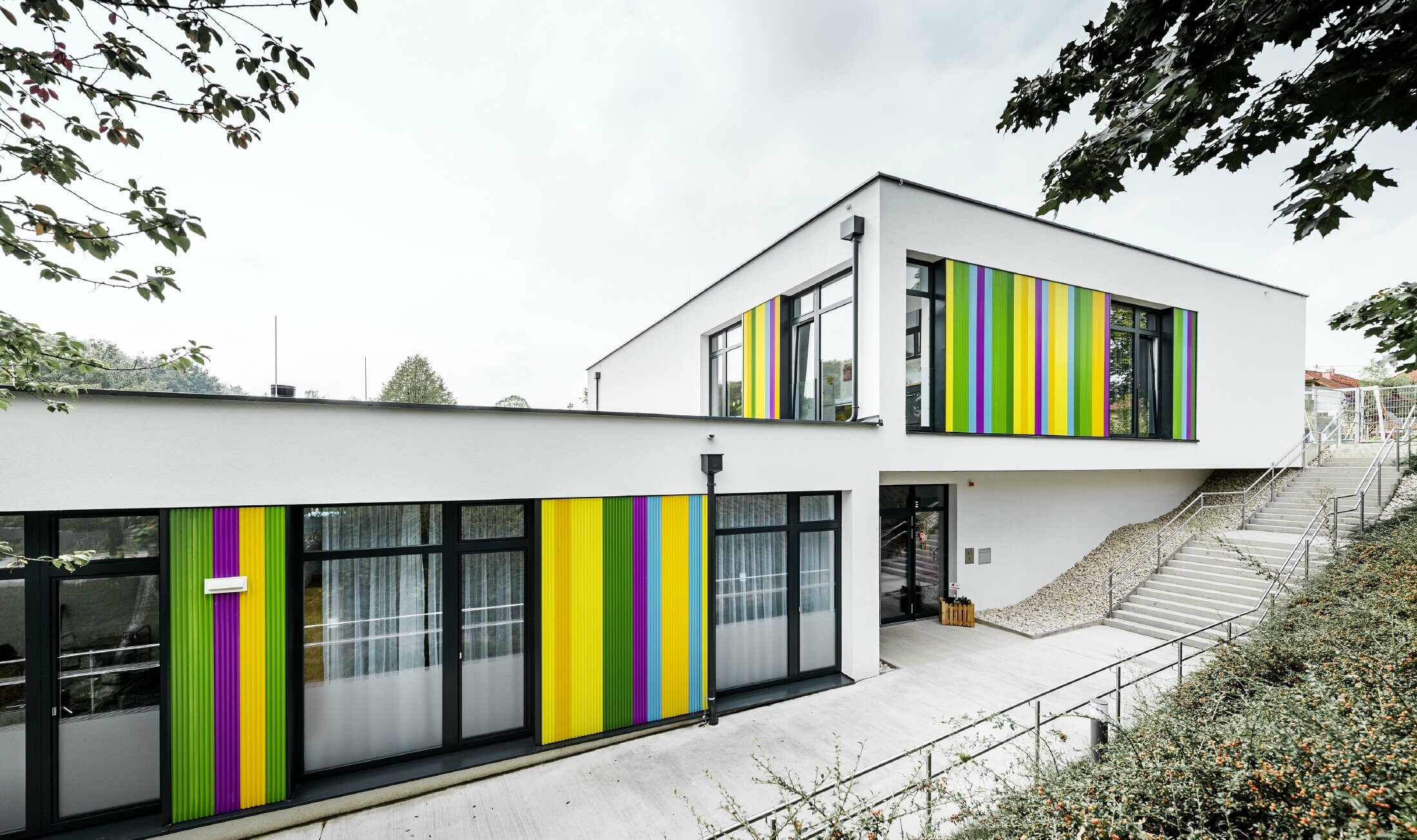 Šareni fasadni elementi tvrtke PREFA za dječji vrtić u Hargelsbergu. Zgrada ima ravni krov i velike prozore od stropa do poda.