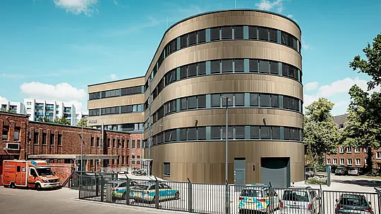 Upečatljiv pogled na Policijsku postaju 43 u Hamburgu otkriva zakrivljenu novogradnju s PREFA aluminijskom fasadom od eloksiranih ekstrudiranih zupčastih profila u srednje brončanoj nijansi. Kao snažan arhitektonski akcent u urbanom prostoru, fasada privlači pažnju svojom finom vertikalnom strukturom koja stvara dinamičnu igru svjetla i sjene te dodatno ističe zaobljenu geometriju objekta. U kontrastu s njom ostaje povijesna opečna struktura postojećeg dijela zgrade. PREFA aluminij pruža izvrsnu otpornost na vremenske utjecaje i koroziju, dug vijek trajanja te minimalno održavanje – idealno rješenje za održive, funkcionalne i estetski zahtjevne fasadne koncepte.
