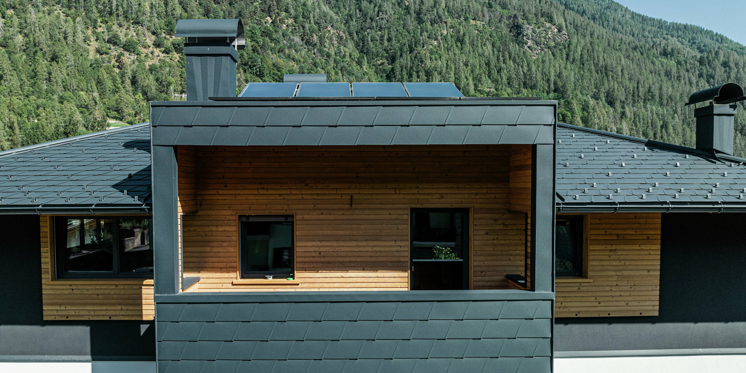 Nahaufnahme der PREFA Dachschindeln DS.19 in P.10 Anthrazit auf dem Dach eines Wohnhauses in Commezzadura, Italien. Die Aluminiumverkleidung sorgt für ein modernes und langlebiges Design am Dach und der Fassade der überdachten Balkone. Die natürliche Holzfassade unter der dunklen Blechverkleidung schafft einen harmonischen Gesamteindruck.