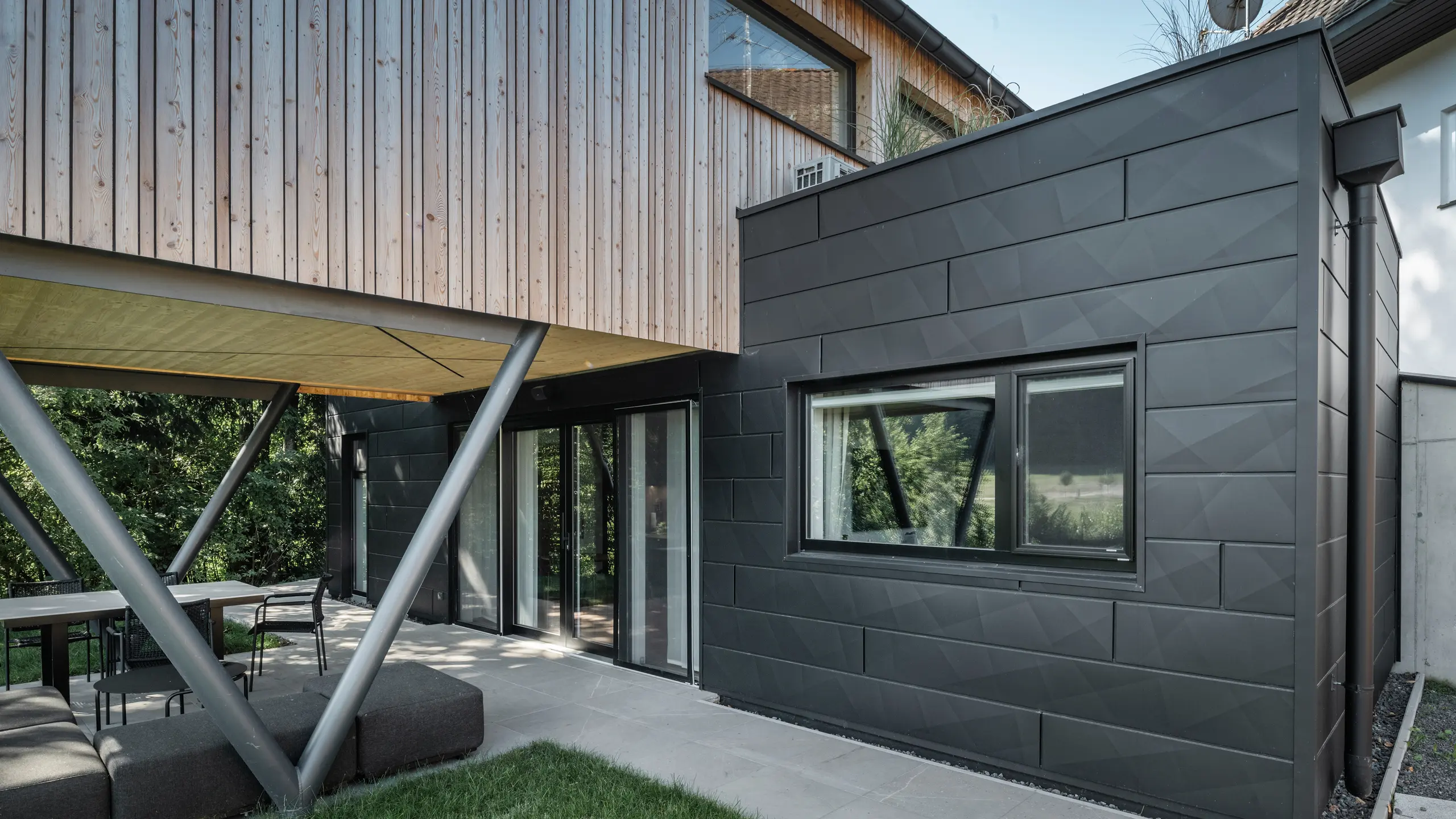 Arhitektonski vrhunac: 'Woodstock Chalet 5' u St. Martin am Innkreis, Gornja Austrija. Moderni vjenčani prostor naglašen je upotrebom antracitno obojenih PREFA Siding.X fasadnih panela. Aluminijski elementi stvaraju upečatljiv kontrast s panelima od svijetlog drva i metalnim potpornim konstrukcijama. Tamni okviri prozora skladno se uklapaju u dizajn pročelja, dajući zgradi elegantan izgled. Ova slika prikazuje estetsku ravnotežu između prirodne teksture drva i industrijskog šarma aluminijskog pročelja, što je reprezentativno za suvremeni dizajn zgrada.