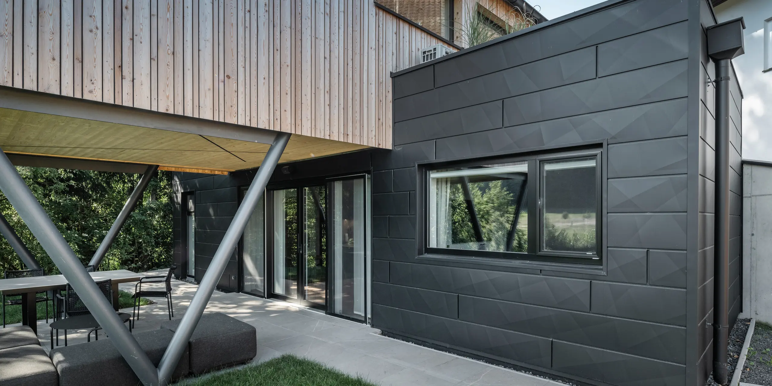 Arhitektonski vrhunac: 'Woodstock Chalet 5' u St. Martin am Innkreis, Gornja Austrija. Moderni vjenčani prostor naglašen je upotrebom antracitno obojenih PREFA Siding.X fasadnih panela. Aluminijski elementi stvaraju upečatljiv kontrast s panelima od svijetlog drva i metalnim potpornim konstrukcijama. Tamni okviri prozora skladno se uklapaju u dizajn pročelja, dajući zgradi elegantan izgled. Ova slika prikazuje estetsku ravnotežu između prirodne teksture drva i industrijskog šarma aluminijskog pročelja, što je reprezentativno za suvremeni dizajn zgrada.