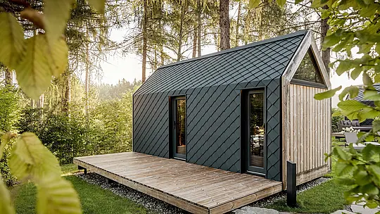 Minimalistička tiny kuća u Secret Koi Gardenu na jezeru Natterer See smještena je u mirnom šumskom krajoliku. Krov i fasada oblikovani su sitnim PREFA krovnim i zidnim rombovima od aluminija u P.10 antracitnoj boji, čija precizna struktura nalik ribljoj koži nježno reflektira sunčevu svjetlost.  Prostrana drvena terasa otvara prostor prema prirodi, dok dugotrajni i vremenski otporni PREFA aluminij štiti arhitekturu i stvara upečatljiv kontrast drvu i zelenilu.