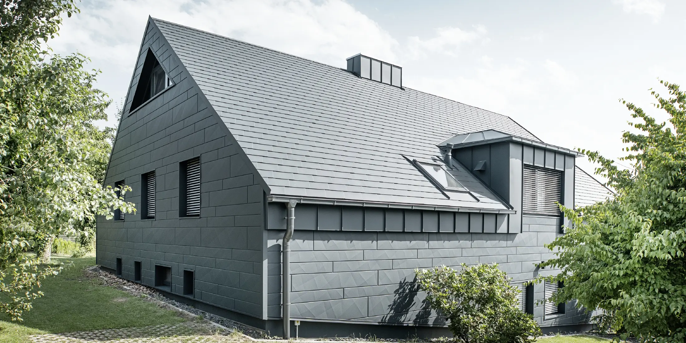 Velika obiteljska kuća s dvovodnim krovom i puno prozora. Cijela kuća obložena je antracit aluminijskom školjkom, na krovu su korištene PREFA aluminijske šindre, a na fasadi Siding.X u antracit boji.