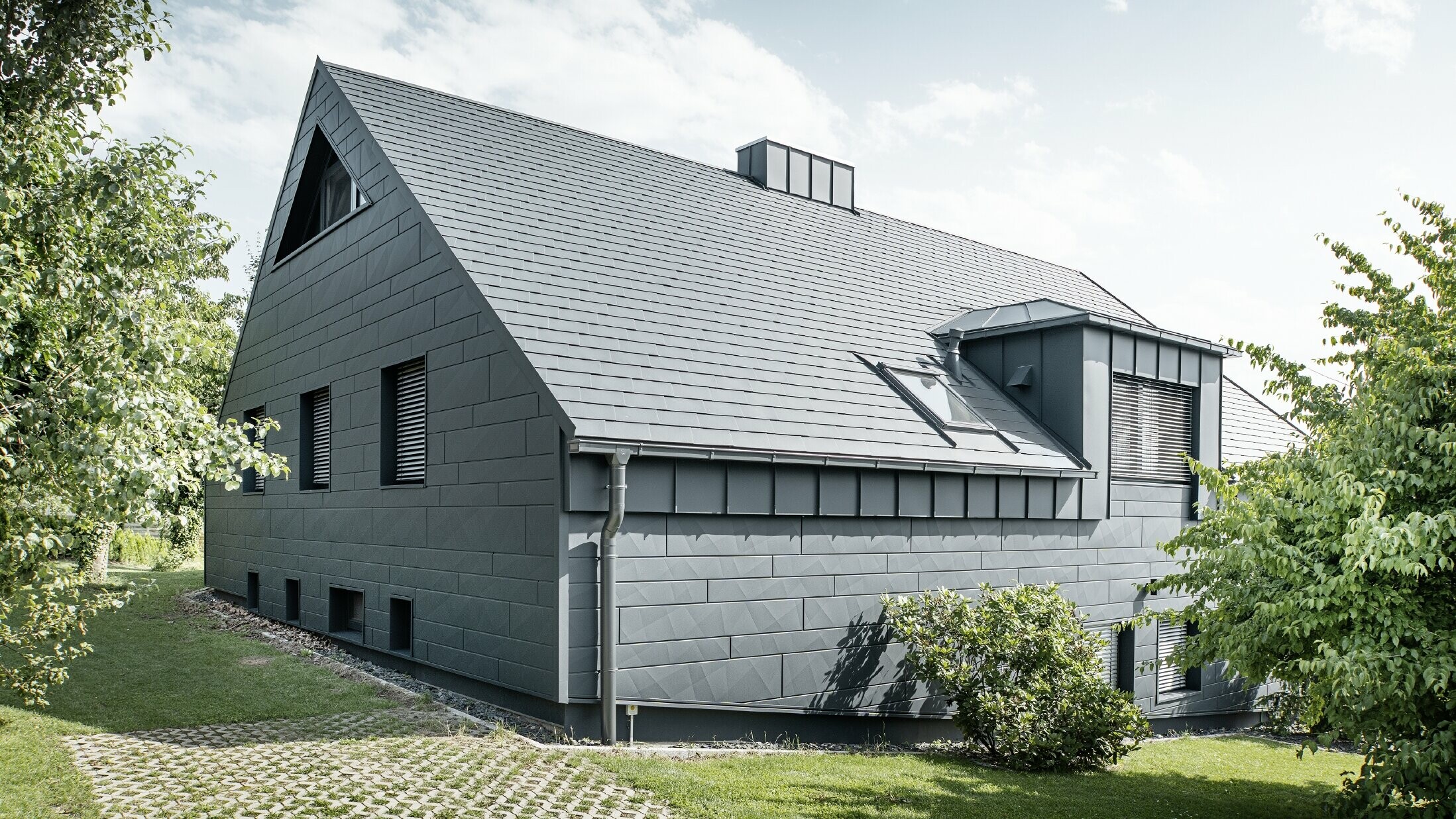Velika obiteljska kuća s dvovodnim krovom i puno prozora. Cijela kuća obložena je antracit aluminijskom školjkom, na krovu su korištene PREFA aluminijske šindre, a na fasadi Siding.X u antracit boji.