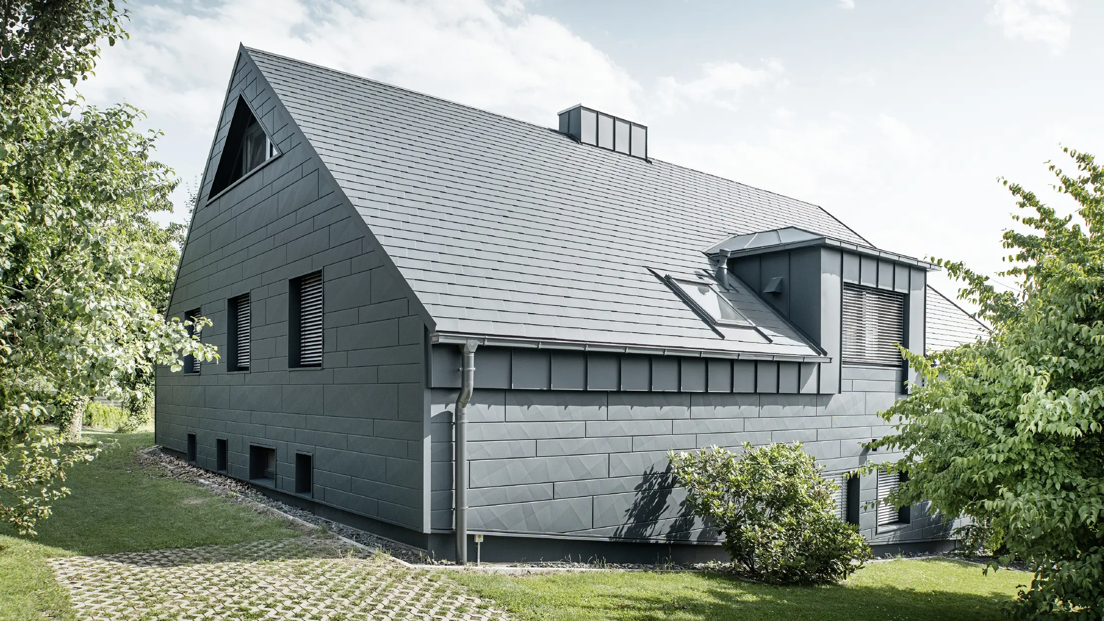 Velika obiteljska kuća s dvovodnim krovom i puno prozora. Cijela kuća obložena je antracit aluminijskom školjkom, na krovu su korištene PREFA aluminijske šindre, a na fasadi Siding.X u antracit boji.