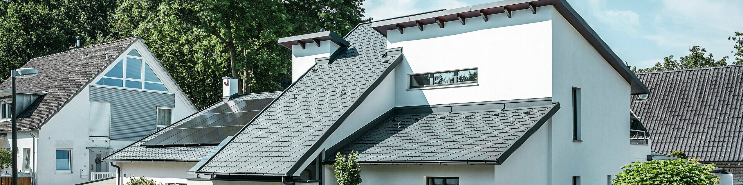 Moderna samostojeća kuća s bijelom žbukanom fasadom i tamnim PREFA aluminijskim krovom s kosim stranama. Na krovu su integrirani fotonaponski moduli i aluminijski oluci u antracitnoj boji. Zgrada se nalazi u dobro održavanom stambenom području s uređenim prednjim vrtovima, popločanim prilazima i okruženom samostojećim kućama.