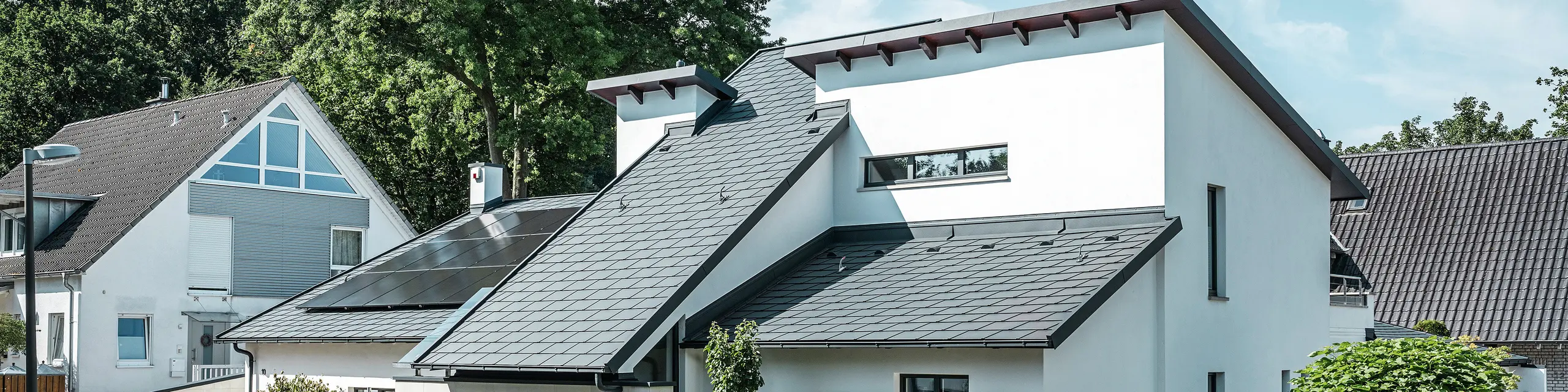 Moderna samostojeća kuća s bijelom žbukanom fasadom i tamnim PREFA aluminijskim krovom s kosim stranama. Na krovu su integrirani fotonaponski moduli i aluminijski oluci u antracitnoj boji. Zgrada se nalazi u dobro održavanom stambenom području s uređenim prednjim vrtovima, popločanim prilazima i okruženom samostojećim kućama.