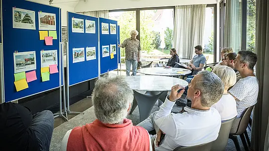 PREFARENZEN 2027 – stručni dijalog u svijetloj, modernoj prostoriji za seminare: Moderator Matthias Finkentey stoji ispred plavih panoa na kojima su predstavljeni arhitektonski projekti s PREFA aluminijskim rješenjima. Žiri sjedi za okruglim stolovima, pažljivo raspravlja i bilježi dojmove. Veliki prozori propuštaju dnevno svjetlo, dok se izvana vidi zeleni vrt. Scena prikazuje povezivanje kreativne arhitektonske rasprave, funkcionalnog planiranja i vrednovanja dizajna. PREFA aluminijski proizvodi predstavljaju dugotrajne, estetski oblikovane krovove i fasade koji u arhitektonskim projektima utjelovljuju inovaciju, kvalitetu i inspiraciju.