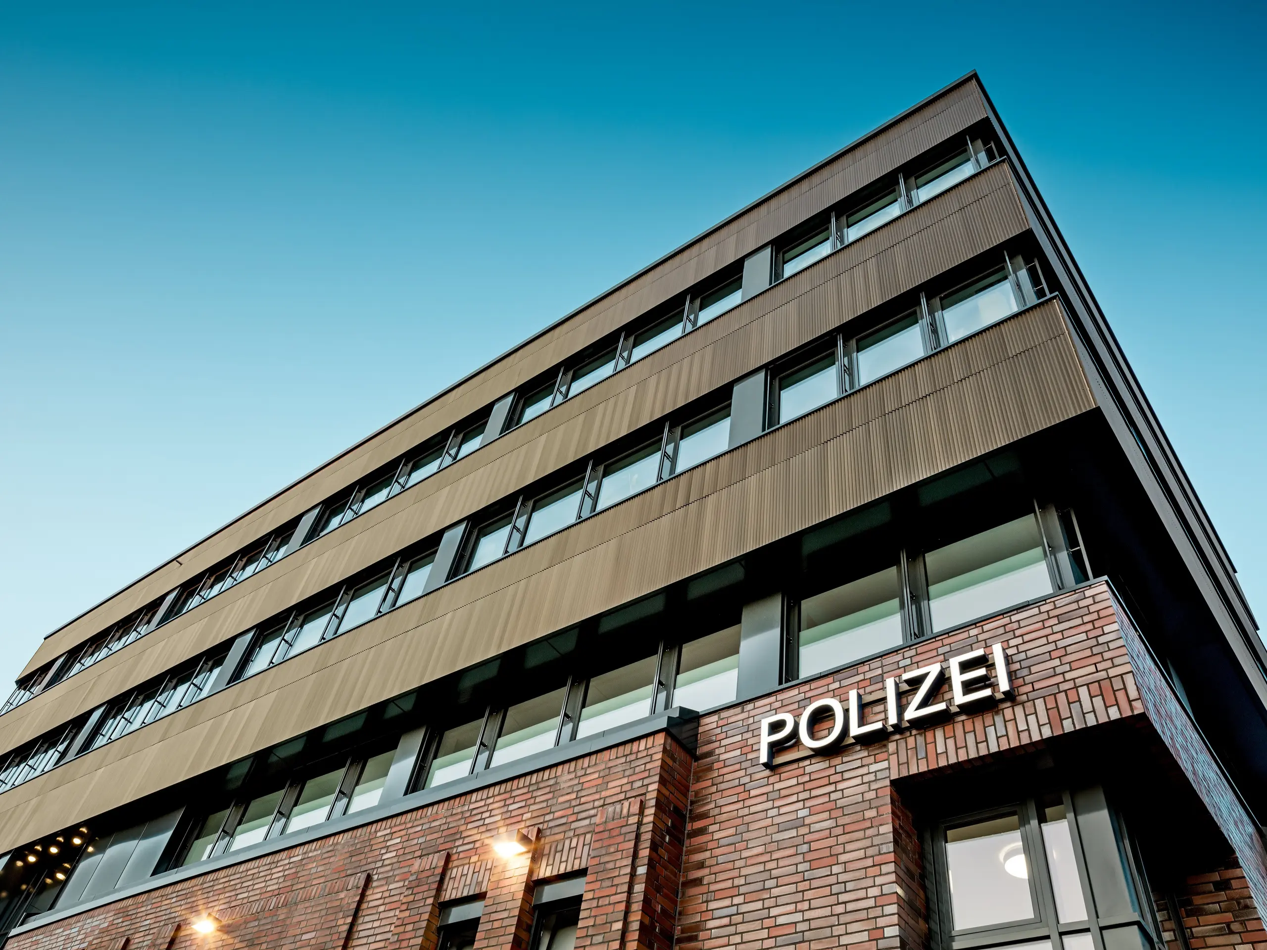 Arhitektonski pogled ispod novog dijela Policijske postaje 43 u Hamburgu otkriva precizno strukturiranu novogradnju koja se izdiže iznad postojeće opečne arhitekture. Posebnu pažnju privlači PREFA aluminijska fasada od ekstrudiranih zupčastih profila u elegantnoj srednje brončanoj nijansi. Fino rebrasta površina djeluje kao snažan vizualni element, stvarajući fascinantnu igru svjetla i refleksija duž jasno oblikovanih horizontalnih prozorskih nizova. PREFA aluminij pruža izvrsnu otpornost na koroziju, dug vijek trajanja i minimalne zahtjeve za održavanje – održivo, funkcionalno i dizajnerski sofisticirano rješenje za suvremenu fasadnu arhitekturu