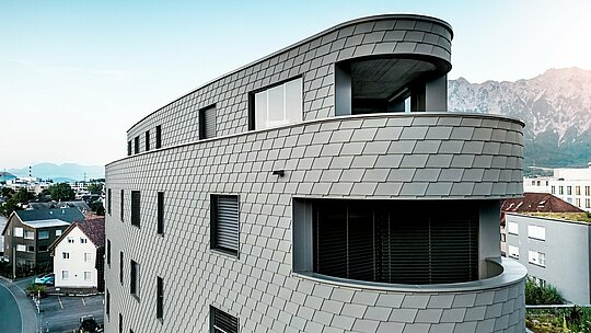 Detaljni kosi prikaz Bernerhausa u Buchsu s nježno zaobljenim kutom i kontinuiranim balkonima. Fasada od PREFA zidnih šindra u P.10 svijetlosivoj boji prekriva trokutastu kubaturu poput precizno obrađene aluminijske kože i stvara živopisni, ljuskasti uzorak. Homogena površina naglašava tekuće radijuse i igru svjetla i sjene. Zahvaljujući laganom, otpornom na vremenske uvjete i dugotrajnom PREFA aluminiju, nastaje gotovo bezdržavna ovojnica zgrade s upečatljivim vizualnim efektom na alpskoj pozadini.