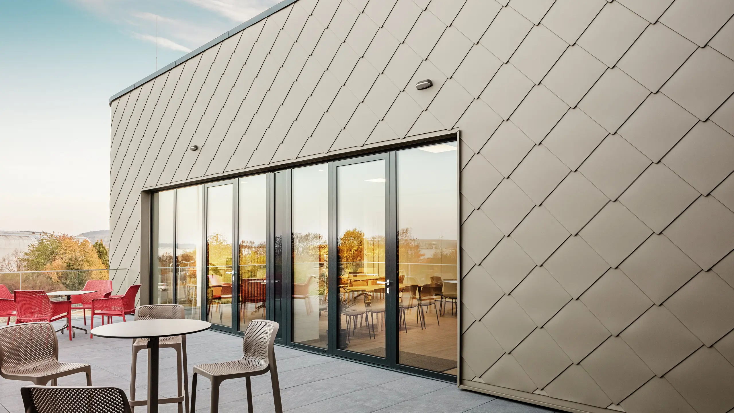 Fasada nove PREFA lokacije u Törökbálintu impresionira svojim elegantnim PREFA zidnim pločicama dimenzija 44 × 44 u boji P.10 Prefabronze. Precizno ugrađena aluminijska fasada stvara moderan, dinamičan izgled uz maksimalnu zaštitu od vremenskih uvjeta. Velikodušna staklena fasada povezuje unutarnji i vanjski prostor te stvara svijetlu i privlačnu atmosferu na prostranoj terasi.