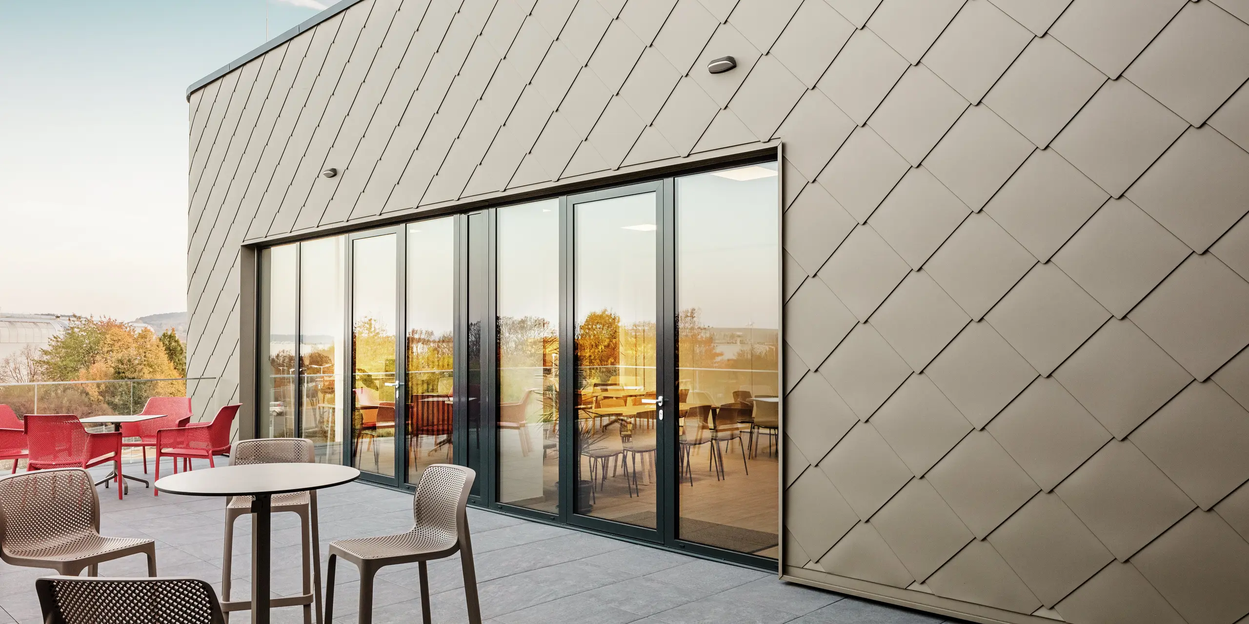 Fasada nove PREFA lokacije u Törökbálintu impresionira svojim elegantnim PREFA zidnim pločicama dimenzija 44 × 44 u boji P.10 Prefabronze. Precizno ugrađena aluminijska fasada stvara moderan, dinamičan izgled uz maksimalnu zaštitu od vremenskih uvjeta. Velikodušna staklena fasada povezuje unutarnji i vanjski prostor te stvara svijetlu i privlačnu atmosferu na prostranoj terasi.