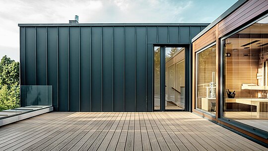 Snimka prikazuje prostranu krovnu terasu budimpeštanske višegeneracijske vile, čija je fasada obložena PREFALZ aluminijem u boji P.10 antracit. Precizni, vertikalno vođeni stojeći falcevi stvaraju jasnu i modernu linijsku strukturu koja se skladno nadovezuje na toplo drvo susjedne saune. Prirodno sunčevo svjetlo dodatno naglašava elegantan kontrast materijala i ističe visokokvalitetnu, dugotrajnu PREFA izvedbu. Prostrana terasa otvara pogled na zelenilo i naglašava suvremeni, komforno orijentirani karakter objekta.