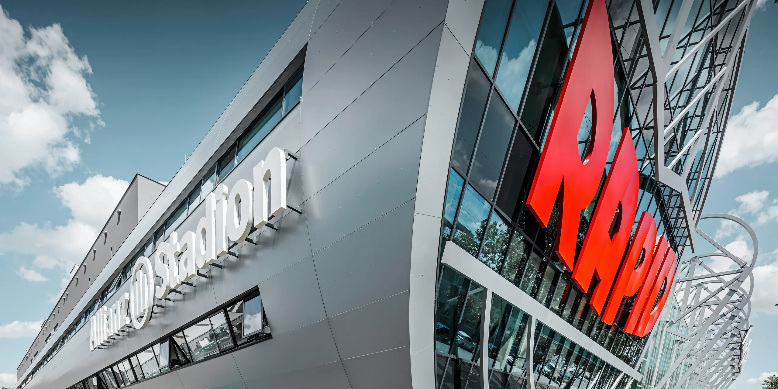 Nahaufnahme der Fassade des Allianz Stadions, Heimat des SK Rapid Wien. Die eindrucksvolle Röhre im Eingangsbereich präsentiert die PREFA Aluminium Verbundplatten in Silbermetallic. Der Blickwinkel unterstreicht die moderne Architektur. Durch den strahlend roten RAPID-Schriftzug und die klare Linienführung der silbernen Platten erhält die Röhre eine visuell beeindruckende Dynamik und Ästhetik. Auf diese Weise wird bereits beim Gang zum Stadion bei allen Fußball-Fans Leidenschaft entfacht.