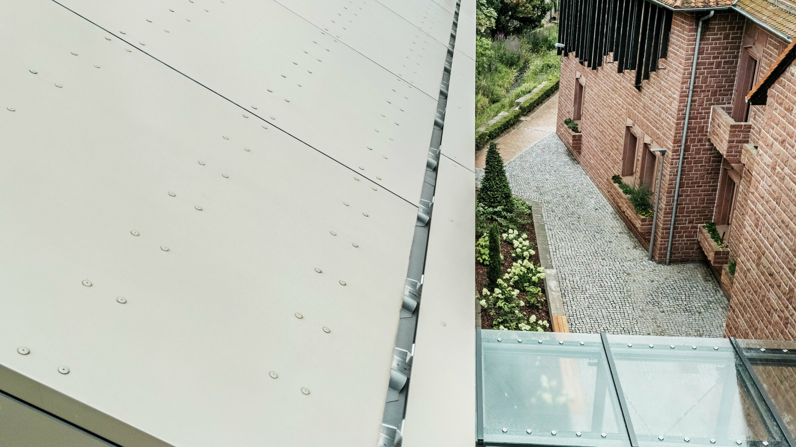 Detaljni prikaz krova gradske vijećnice u Alzenauu, prekrivenog PREFA aluminijskim kompozitnim pločama u nježnoj brončanoj nijansi. Ploče su precizno postavljene i opremljene vidljivim elementima pričvršćivanja, što fasadi daje moderan, industrijski izgled. U prvom planu vidi se stakleni spojni hodnik koji povezuje vijećnicu s obližnjom zgradom.