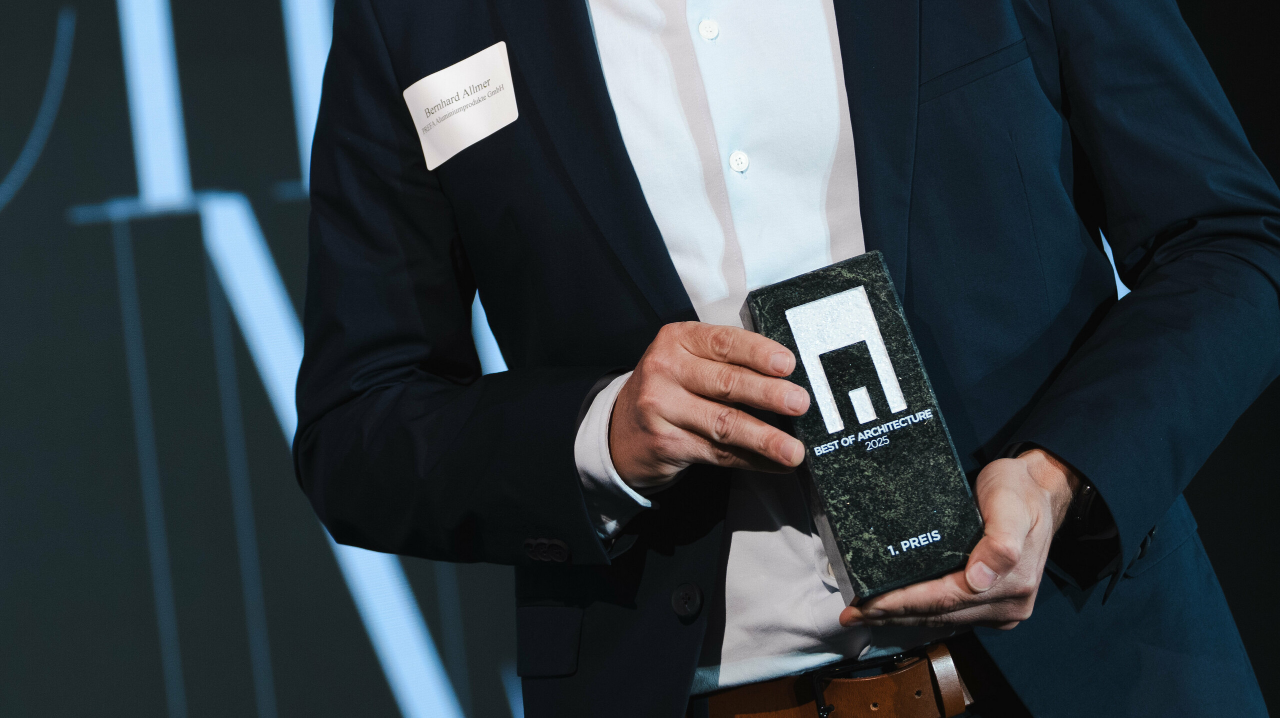 Dodjela nagrade na renomiranom Callwey natječaju „Stambene zgrade godine“ 2025.: u prvom planu elegantna osoba u tamnom odijelu i bijeloj košulji ponosno predstavlja nagradu „Architects’ Choice“. Osoba je Bernhard Allmer, odgovoran za PREFA solarne proizvode. Nagrada se sastoji od elegantne tamne kamene ploče s modernim logom i čistom tipografijom. U pozadini je stilska pozornica s velikim natpisom i naglašenim osvjetljenjem, što naglašava svečani karakter događaja. Scena odražava inovaciju i izvrsnost u arhitekturi.