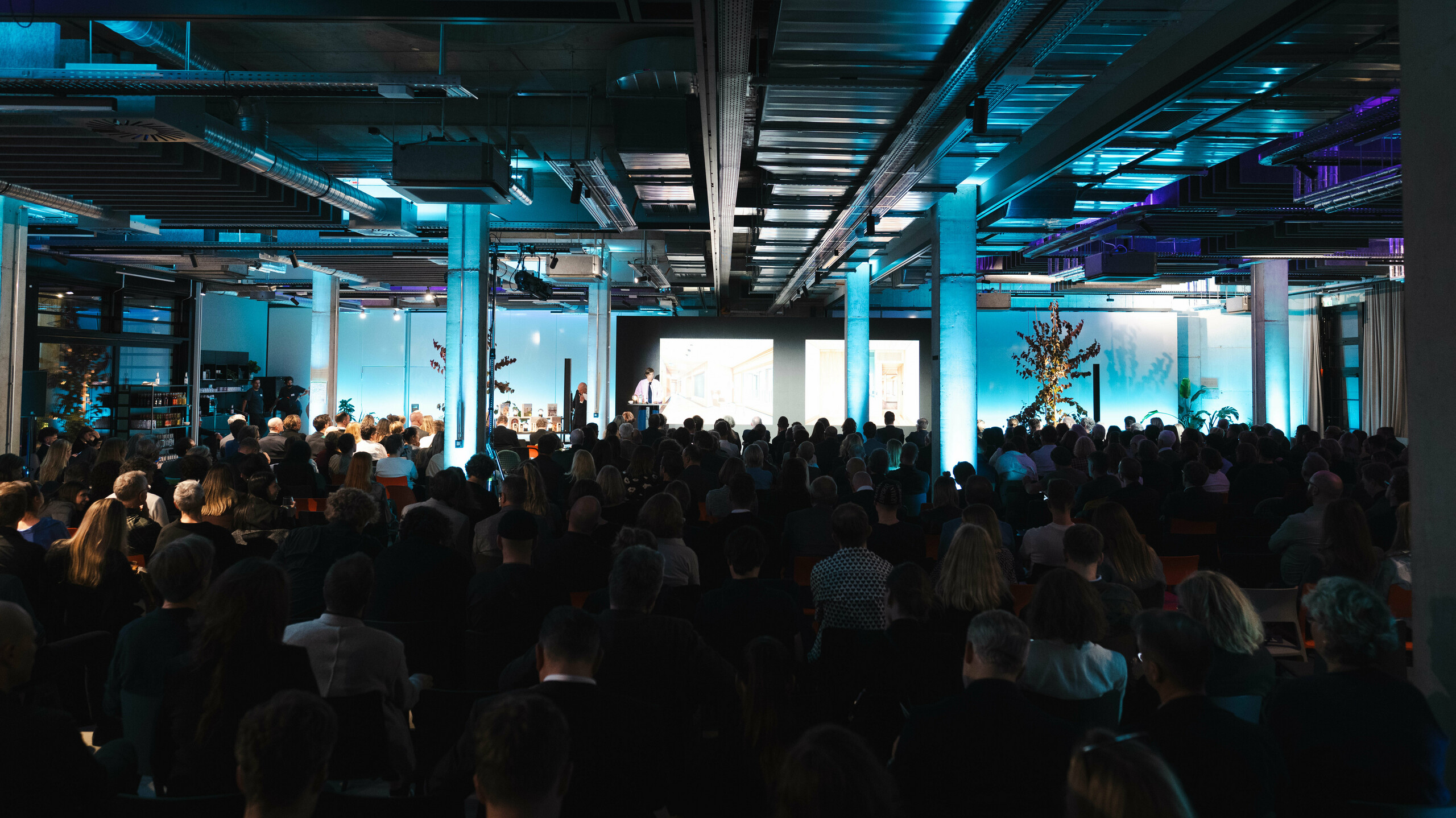 Atmosferična scena s dodjele nagrade "Stambena zgrada godine 2025": pogled na prepunu dvoranu u modernoj industrijskoj zgradi s upečatljivim plavim osvjetljenjem. U prvom planu, pozornica s velikim zaslonom prikazuje prezentacije. Arhitektura prostorije, sa svojim izloženim čeličnim gredama i betonskim stupovima, naglašava inovativni karakter događaja koji slavi održiva rješenja poput solarnog modula PREFALZ.