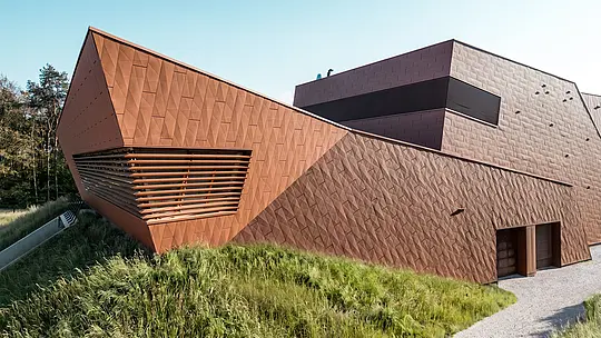 Izražajna crpna stanica za pitku vodu WML u Lottumu s dinamično presavijenim volumenom zgrade i upečatljivim vrhom kao vizualnim atraktivnim elementom. PREFA Siding.X aluminijska fasada u toplom izgledu hrđe impresionira svojom fasetiranom, živopisnom strukturom i preciznom igrom svjetla i sjene. Horizontalne lamele naglašavaju dubinu arhitekture, dok se zgrada skladno uklapa u prirodno okruženje. Lagani, dugotrajni i reciklabilni PREFA aluminijski paneli predstavljaju vrhunsku otpornost na vremenske uvjete te održiv fasadni omotač koji zahtijeva minimalno održavanje.