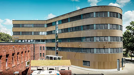Dinamičan pogled na vanjski izgled proširene Policijske postaje 43 u Hamburgu otkriva zakrivljenu novogradnju s približno 1.100 m² PREFA aluminijske fasade izvedene od ekstrudiranih zupčastih profila u srednje brončanoj eloksiranoj nijansi. Kao upečatljiv arhitektonski element, fasada stvara snažan kontrast postojećoj opečnoj strukturi. Fino oblikovana površina mijenja se kroz igru svjetla i sjene, dajući fasadi dodatnu dubinu i precizno definiran karakter. Zahvaljujući izvrsnoj otpornosti na koroziju, dugom vijeku trajanja i minimalnim zahtjevima za održavanje, PREFA aluminijska rješenja omogućuju održivu, funkcionalnu i arhitektonski sofisticiranu kvalitetu gradnje.