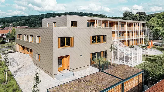 Iz povišene perspektive slika prikazuje L‑oblik novog objekta dječjeg vrtića u Alzenauu s pripadajućim stambenim dijelom – budućnosno orijentiranu hibridnu zgradu izgrađenu održivo. Prednja ventilirana PREFA aluminijska fasada u brončanoj boji s zidnim rombovima 44 × 44 oblikuje modernu, strukturiranu vanjsku „odoru“ vrtićkog dijela. Prostrani drveni prozori i vertikalne lamelice stvaraju prijateljski, topao izgled. Stražnji prostor s vanjskim stubištem, igralištima i zelenim površinama ilustrira funkcionalnu odvojenost, ali i povezivanje različitih namjena. Zeleni krov te kombinacija materijala – aluminija, drva i betona – odražavaju visoke ekološke standarde projekta, čineći snažnu poruku održive obrazovne i stambene arhitekture.