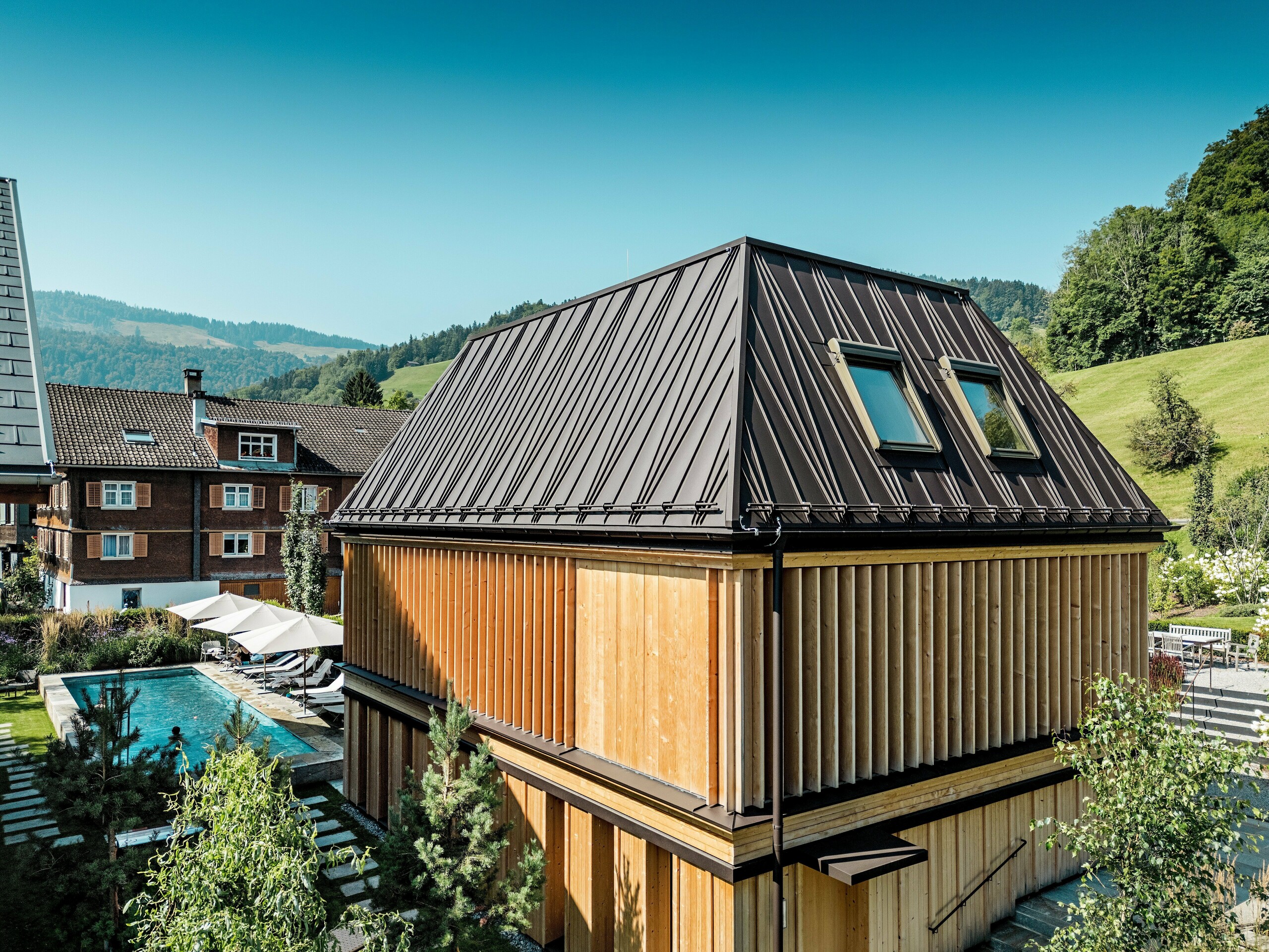 Moderni „Badehaus“ hotela Hirschen u Schwarzenbergu na sunčan dan, smješten u zelenom brežuljkastom krajoliku Bregenzerwalda. Zgrada impresionira svojim upečatljivim PREFA krovom s falcom u boji P.10 orah smeđa s izraženim, dijagonalno postavljenim falc uzorkom i dva krovna prozora. Fasada se sastoji od etažom visokih, vertikalnih drvenih lamela koje fleksibilno upravljaju svjetlom, sjenom i privatnošću. U lijevom dijelu slike vidi se vanjski bazen s prostorom za ležaljke – mjesto mira i opuštanja. Spoj visokokvalitetne arhitekture, regionalnih materijala i promišljene izvedbe skladno se uklapa u tradicionalno okruženje.