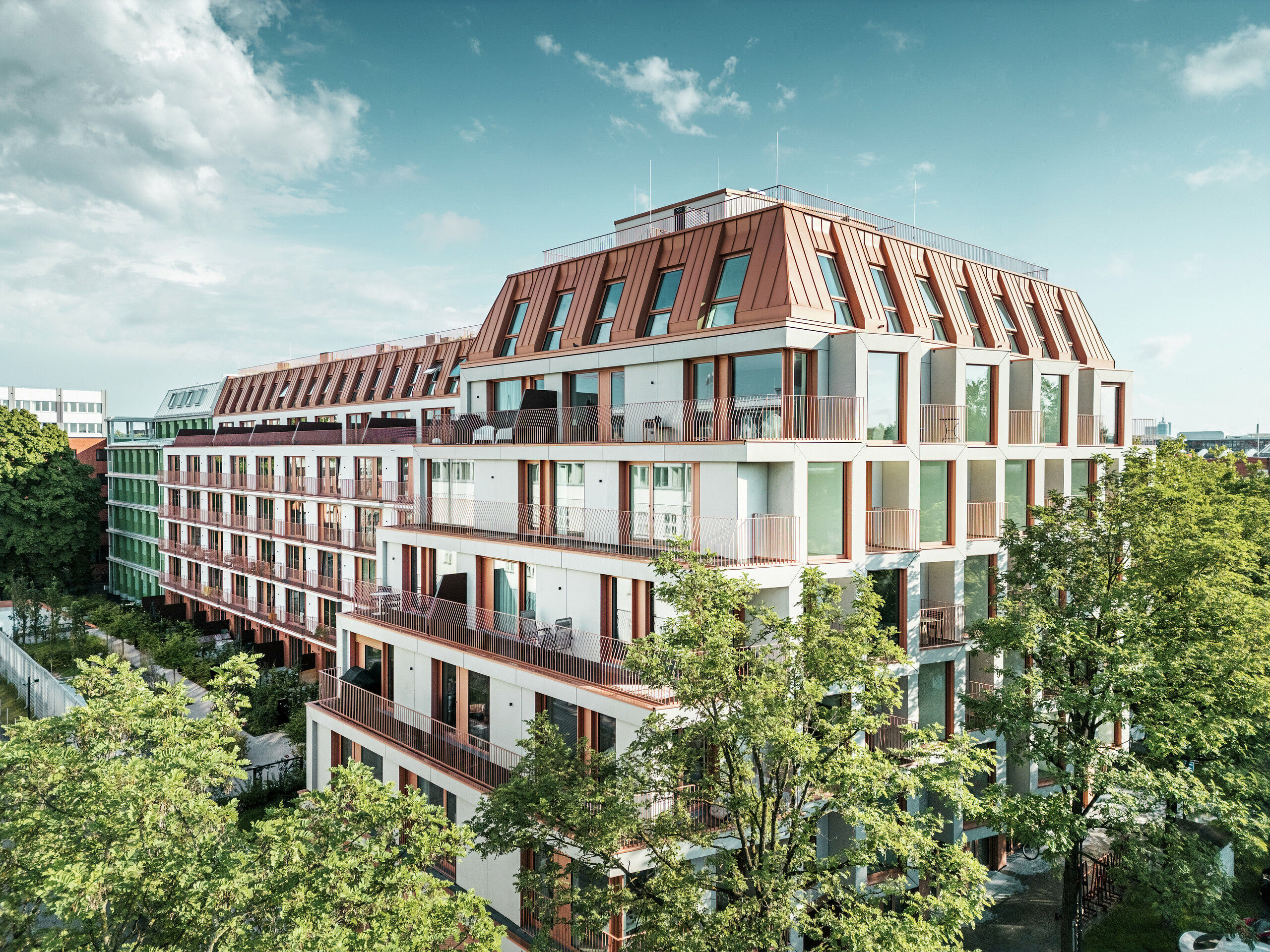 Cjelokupni pogled na urbani stambeni projekt Van B u Münchenu s jasnom arhitekturom, zelenim vanjskim prostorima i upečatljivim PREFALZ aluminijskim krovom u boji IGP Orange HDP tvrtke PREFA. Otporno i malo održavano krovno materijalo čini povezujući dizajnerski element zgrade i osigurava dugotrajno, estetsko rješenje u urbanom prostoru. Prostrane staklene površine, balkoni i promišljeni tlocrt nude suvremeni stambeni komfor visoke dizajnerske kvalitete.