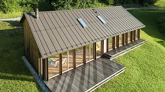 Moderna drvena kuća s prostranom staklenom fasadom i izdržljivim PREFALZ aluminijskim krovom u boji P.10 smeđa – idealna za padinske terene poput onih u Vinju. Kvalitetni krovni pokrov osigurava maksimalnu otpornost na vremenske uvjete, dug vijek trajanja i elegantan izgled. Precizno postavljeni stojeći falcovi i integrirani krovni prozori stvaraju prozračan i održiv stambeni koncept. Pogled iz zraka otkriva preciznu obradu aluminijskih stojećih falcova.