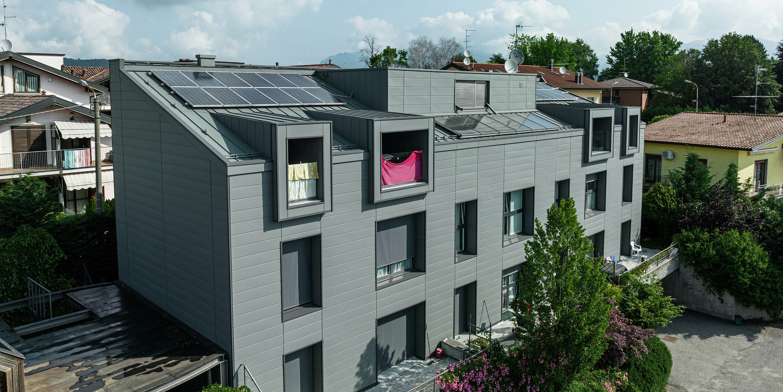 Blick von oben auf die moderne Wohnanlage in Faloppio, Italien, mit einer eleganten PREFA Siding-Fassade und einem mit PV-Modulen bestückten Dach. Die langlebige Aluminiumverkleidung, die umweltfreundliche Energiegewinnung durch die Kraft der Sonne und die moderne Optik ergeben ein harmonisches Gesamtpaket. Die Kombination aus ästhetischer Architektur und nachhaltiger Technologie macht das Gebäude zu einem Paradebeispiel für zeitgemäßen Wohnbau.