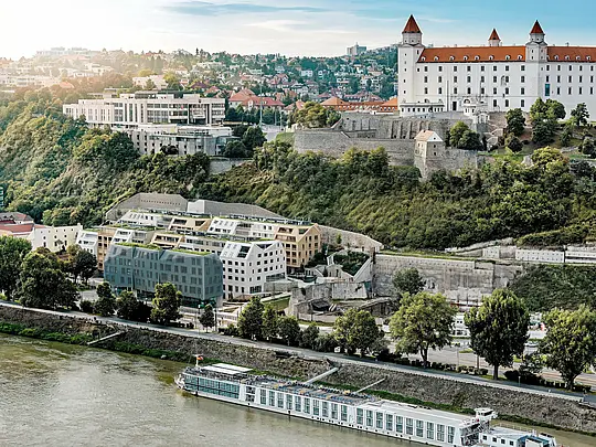 Pogled s visine na četvrt Vydrica u Bratislavi, s pogledom na dvorac i Dunav: slojevita arhitektura oduševljava zelenim krovnim površinama, strmim dvostrešnim krovovima i precizno uklopljenim krovnim prozorima kao vizualnim naglascima. PREFA aluminijske krovne šindre u bojama Prefa bijela, pješčano smeđa i brončana povezuju građevinske cjeline u skladnu cjelinu te se uklapaju u povijesno okruženje. Dugotrajna, lagana i iznimno otporna PREFA rješenja omogućuju izvedbu složenih krovnih geometrija i osiguravaju održivu, jednostavnu za održavanje građevinsku ovojnicu visoke estetske kvalitete.