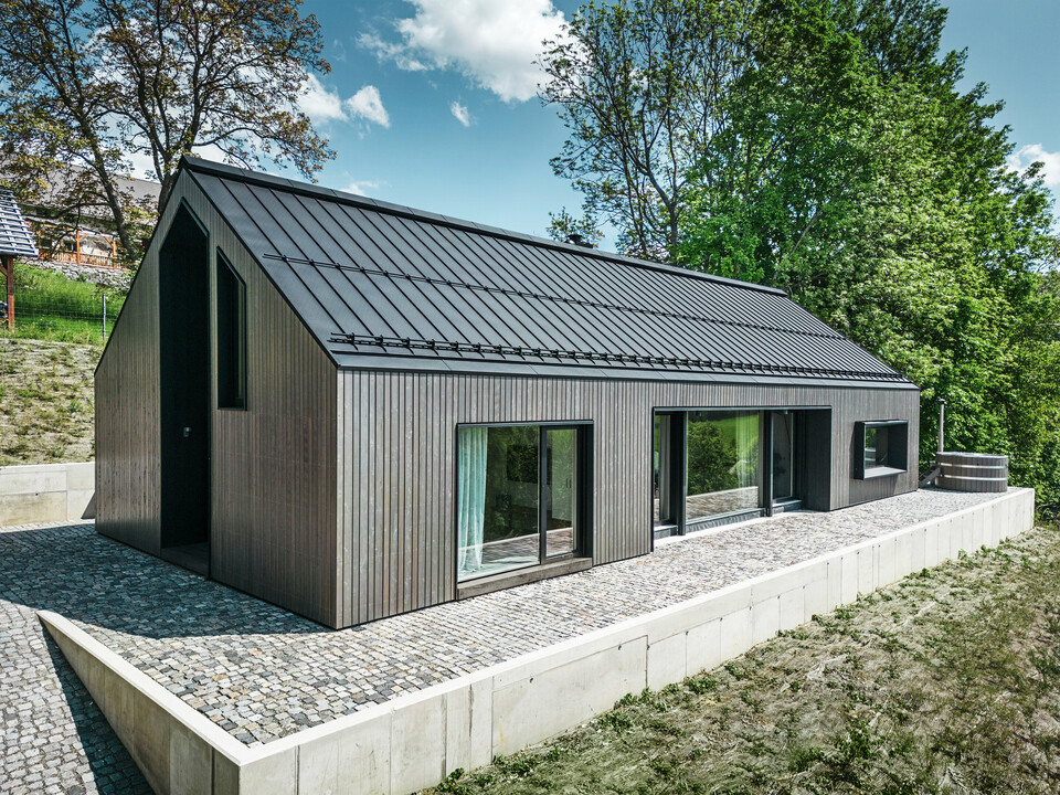 Vorderseite eines Holzmassivhauses in Kuncice, CZ. Das langlebige Aluminiumdach besteht aus PREFALZ in elegantem Schwarz. Das Haus verfügt über großzügige Fensterflächen und einen Whirlpool im Außenbereich.