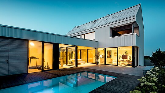 Slika prikazuje modernu obiteljsku kuću u Freistadtu s upečatljivom PREFALZ-fasadom u čistobijeloj boji. Precizno naborana aluminijska fasada daje zgradi jasnu, geometrijsku eleganciju koja se skladno povezuje s prostranim prozorima i ravnim krovom. Korištenje PREFALZ-a osigurava ne samo estetsku postojanost, već i visoku otpornost na vremenske uvjete te dugovječnost. Otvoreni unutarnji prostor s pogledom na bazen savršeno je upotpunjen čistim linijama i minimalističkim dizajnom, čime kuća predstavlja suvremeno, održivo rješenje za život.