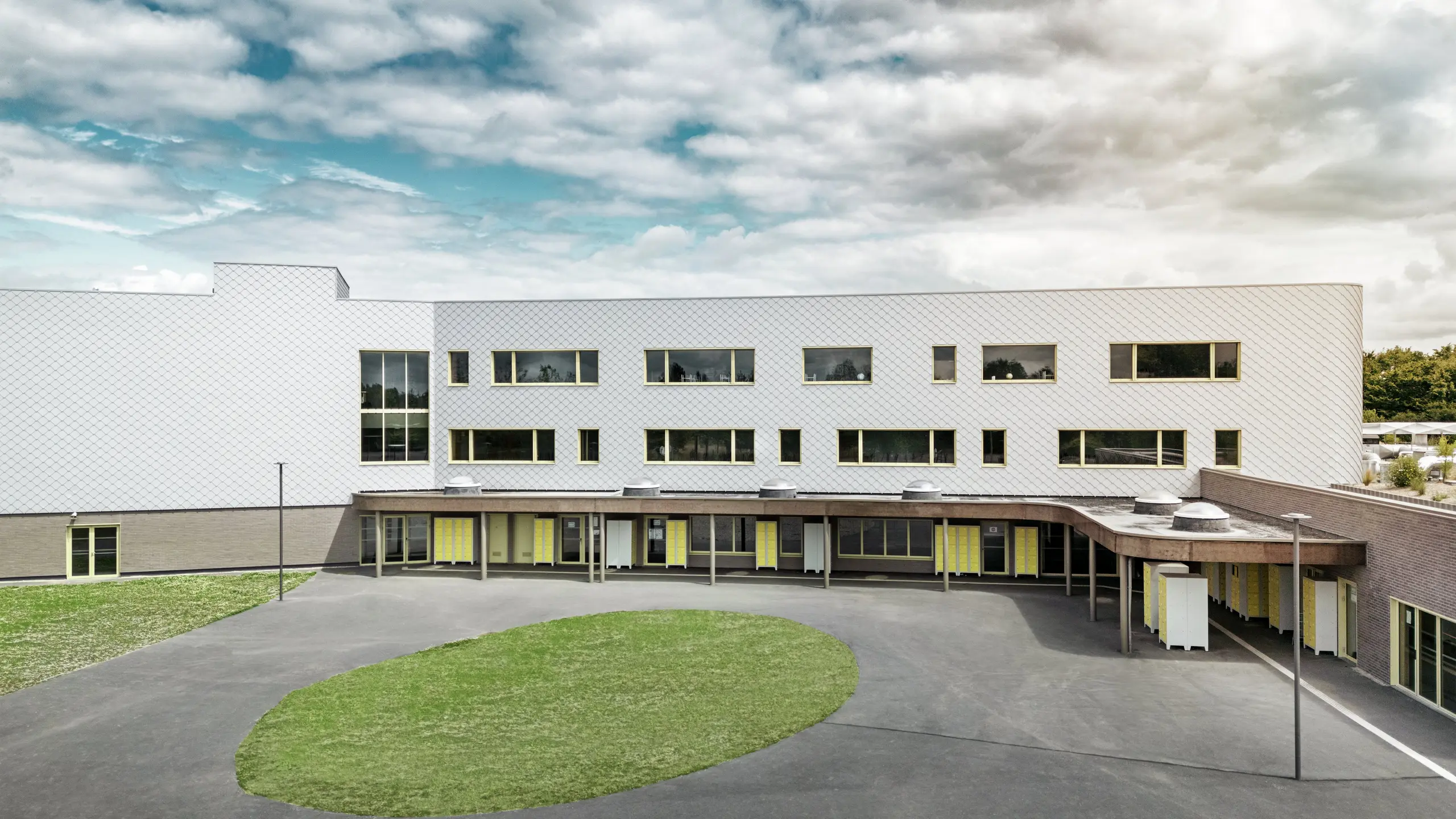 Stražnja strana Collège Léontine Dolivet s prostranim školskim dvorištem i jasnom arhitekturom. Fasada je izrađena od srebrno-metalnih PREFA zidnih rombova, što zgradi daje moderan i elegantan izgled. Više redova prozora i žuta vrata razbijaju strukturu, dok asfaltirano dvorište s centralnom zelenom površinom pruža dovoljno prostora za odmore. Dugotrajna aluminijska fasada kombinira lakoću, otpornost na vremenske uvjete i dizajn – funkcionalan i estetski privlačan objekt u Cesson-Sévignéu, blizu Rennesa.