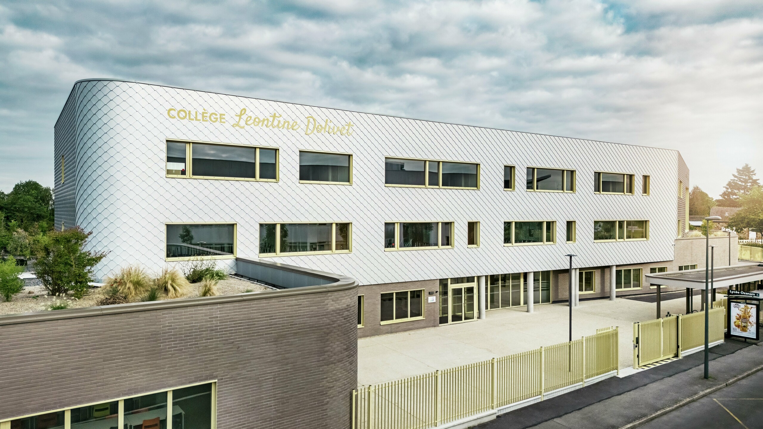 Pogled s prednje strane Collège Léontine Dolivet u Cesson-Sévignéu prikazuje modernu aluminijsku fasadu od PREFA zidnih rombova u srebrno-metalnoj boji. Promjenjiva površina zgradi daje osjećaj lakoće, dok jasne linije i veliki prozori osiguravaju prozirnost. Žuti okviri prozora stvaraju živopisne naglaske. U prvom planu vidljivi su svijetli predprostor, ograda i susjedna ulica. Dugotrajno PREFA rješenje fasade spaja dizajn, otpornost na vremenske uvjete i održivost – arhitektonski privlačan objekt s elegantnim oblikovnim jezikom.