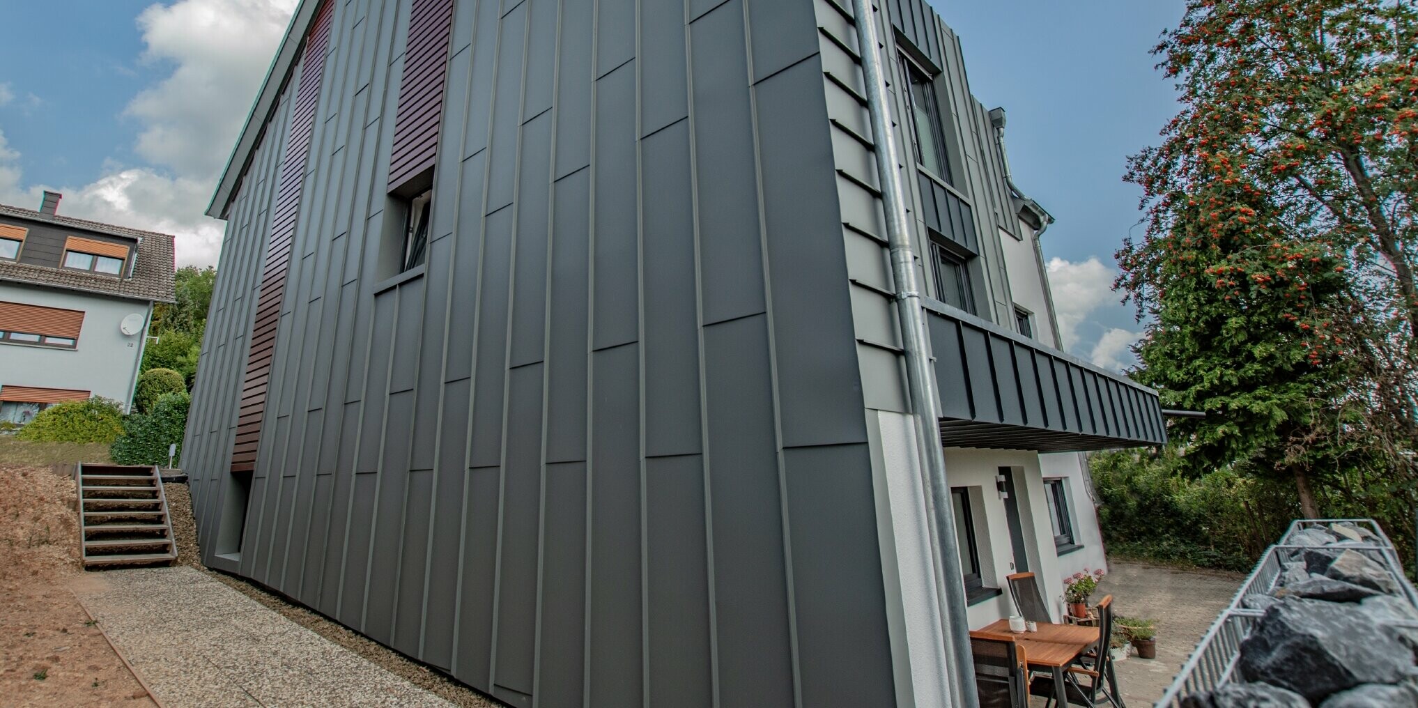 Oblikovanje fasade s vertikalnom fasadnom oblogom od kutnog stojećeg falca u svijetlo sivoj i fasadnim panelima, horizontalno položenim u boji tamnog drva. Kompletna fasada izrađena je od aluminija tvrtke PREFA.