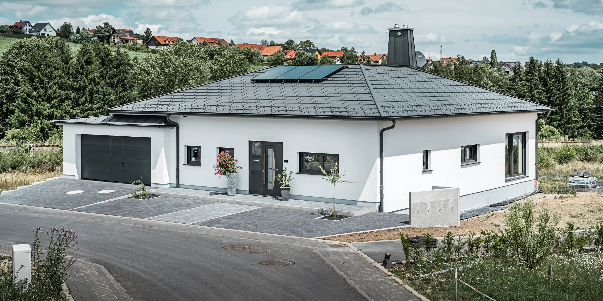 Elegantna samostojeća kuća u Ilmenauu, Turingija, s visokokvalitetnim PREFA krovna ploča u boji P.10 tamnosiva, opremljena robusnim R.16 krovnim pločama. Garaža je u boji i stilu usklađena zahvaljujući upotrebi PREFALZ-a. Upadljive značajke nekretnine su funkcionalno dizajnirani atrij i dimnjak ukrašen PREFA bikom, koji naglašavaju moderan dizajn i inovativnu krovnu tehnologiju tvrtke PREFA. Solarni paneli besprijekorno se uklapaju u održivi koncept kuće. Dobro promišljen cjelokupni koncept bungalova zaokružuje PREFA sustav za odvodnju krova, koji je također u tamnosivoj boji.